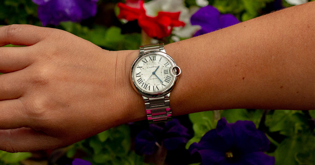 A Cartier Ballon Bleu watch featured on a ladies wrist with purple and red flowers in the background.
